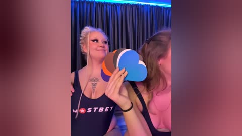 Media: Video of a blonde woman with a tattooed chest, wearing a \"MostBet\" tank top, holding a blue and orange heart-shaped microphone in a dimly lit room with dark curtains.