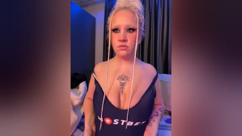 Media: Video of a fair-skinned woman with platinum blonde hair styled in a high ponytail, wearing a low-cut, navy blue tank top revealing ample cleavage and a tattoo on her chest. She stands indoors with dark curtains in the background.