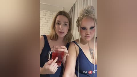 Media: Video of two young women with fair skin, blonde hair, and blue eyes, wearing dark blue tops, standing in a tiled bathroom. One holds a red cup, while the other wears a necklace.