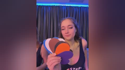 Media: Video of a young woman with light skin, long blonde hair, wearing a black tank top, holding a colorful paper hat with orange, white, and purple stripes, against a blue-lit background with dark curtains.