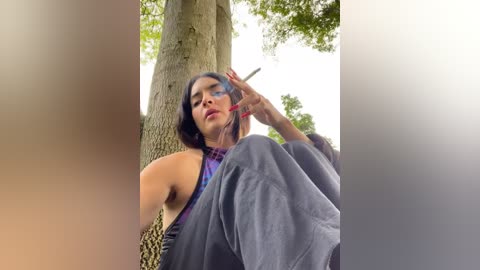 Media: A video of a woman with dark hair and fair skin, wearing a purple top and gray pants, sitting under a tree, holding a lipstick.