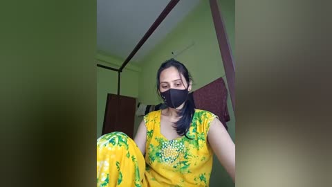 Media: Video of a woman in a yellow and green saree with a black face mask, standing indoors with green walls and wooden furniture.