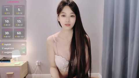 Media: Video of a young Asian woman with long black hair, wearing a pink lace bra, in a minimalistic room with a digital calendar and gray curtains.