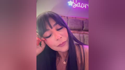 Media: Video of a young woman with long black hair, wearing a black top, resting her head on her hand, with a purple neon sign \"Dance\" in the background.