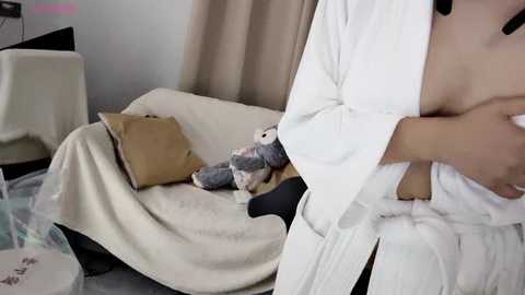 Media: A video of a partially visible person in a white robe, standing in a cozy room with beige curtains, a beige throw, and a plush toy on a beige sofa.