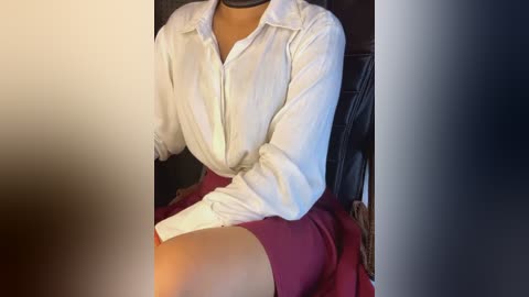 Media: A video of a woman sitting, wearing a white button-down shirt, maroon skirt, and black stockings, with a black choker necklace. She sits in a dark leather chair.