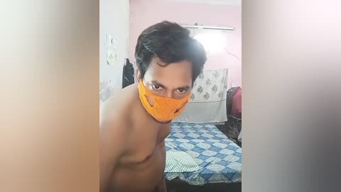 Media: Video of a shirtless man with medium skin tone and short dark hair, wearing an orange face mask, standing in a cluttered room with pastel pink walls, a bed with a blue and white patterned quilt, and a white wall-mounted fan.