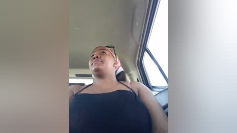 Media: Video of a young woman with light brown skin, wearing a black spaghetti-strap tank top, sitting in a car with the window down, looking up and smiling.