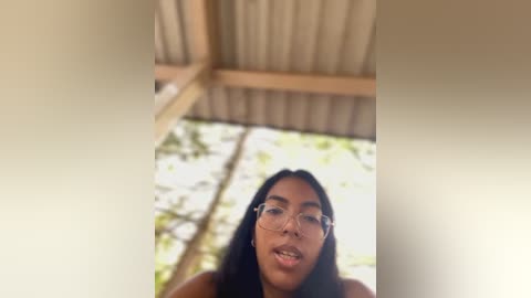 Media: A video of a young woman with medium brown skin, straight black hair, and large glasses, standing in a sunlit, open-air space with wooden beams overhead. The background features blurred greenery and a light-colored floor.