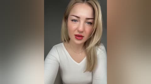 Media: Video of a young Caucasian woman with straight blonde hair, wearing a white ribbed long-sleeve top, red lipstick, and a delicate gold necklace.