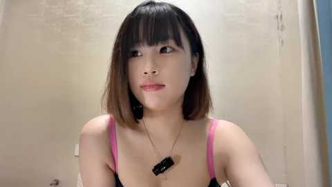 Media: Video of an East Asian woman with straight, shoulder-length brown hair, wearing a pink bra and a black necklace, in a beige tiled bathroom.