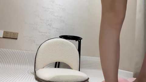 Media: Video of a nude person standing in a modern, minimalist bathroom with white walls, a striped rug, and a textured white chair with black legs.