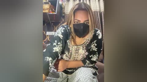 Media: Video of a light-skinned woman with long, straight blonde hair, wearing a black floral blouse, black mask, and black pants, sitting in a cluttered room with a basket and a woven rug in the background.