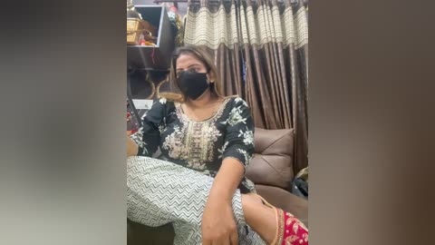 Media: Video of a woman with a black face mask, wearing a black and white floral top and white pants, seated in a living room with brown curtains and a beige couch.