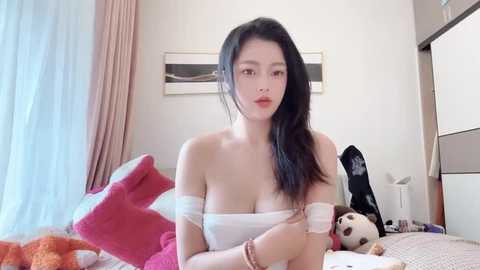 Media: Video of an East Asian woman with long black hair, fair skin, and medium breasts, wearing a white off-shoulder top, sitting on a bed with pink and red pillows, holding a glass of water, in a modern, minimalistic bedroom.