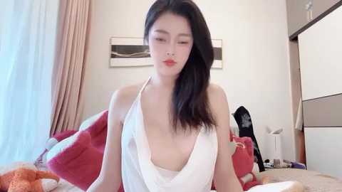 Media: Video of an Asian woman with long black hair, wearing a revealing white halter top, sitting on a bed with red and orange blankets, in a modern bedroom with light curtains and minimalistic decor.