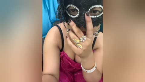 Media: A video of a woman in a black, sequined masquerade mask, with dark hair partially covered, wearing a revealing pink sequined dress, silver bracelets, and large rings.