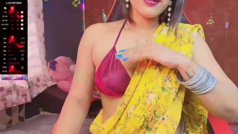 Media: Video of a woman with medium brown skin and dark hair, wearing a yellow sari with a magenta bralette, and multiple silver bangles. Background shows a bed with pink pillows and a colorful room.