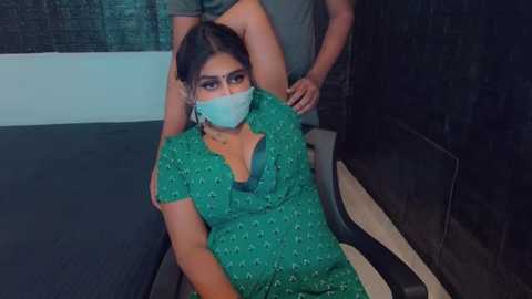 Media: Video of a South Asian woman with medium skin tone, wearing a green saree with white floral patterns, a blue face mask, and a pendant necklace. She sits on a black chair, while a man with light skin stands behind her.