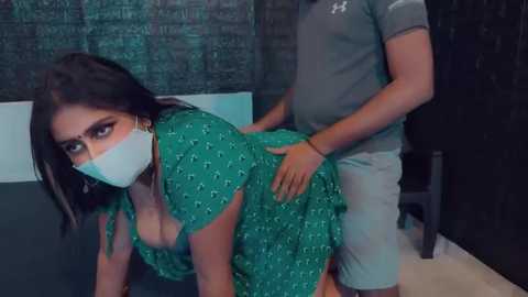 Media: Video of a woman with long dark hair in a green dress, gagged with a white mask, being groped by a man in a gray shirt and shorts in a dimly lit room.