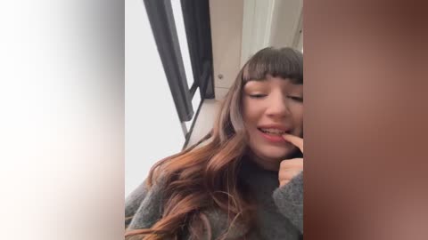 Media: A video of a young woman with long, wavy brown hair, wearing a gray sweater, biting her finger, smiling, taken in a dimly lit room with a white door and black curtains.