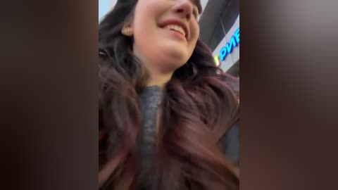 Media: A video captures a close-up of a smiling woman with long, dark hair, wearing a dark jacket. The background features blurred store signage in blue and green.