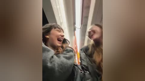 Media: Video of two laughing women on a subway train, one with long brown hair, one with blonde hair, wearing dark coats, laughing hysterically, orange and white subway poles in background.