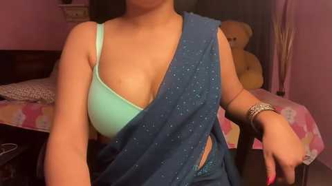 Media: Video of a woman with medium skin tone wearing a light blue bra and a blue saree with white speckles, in a pink bedroom with a teddy bear and a floral-patterned bedspread.