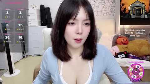 Media: Video of a young Asian woman with shoulder-length black hair, wearing a light blue cardigan over a white lace bra, seated in a cozy, cluttered room with a TV displaying a video game.