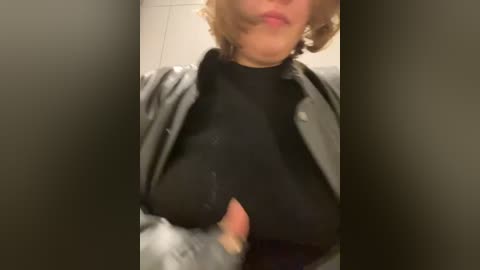 Media: Video of a woman with short, curly hair in a black turtleneck and grey leather jacket, partially exposing her breasts.