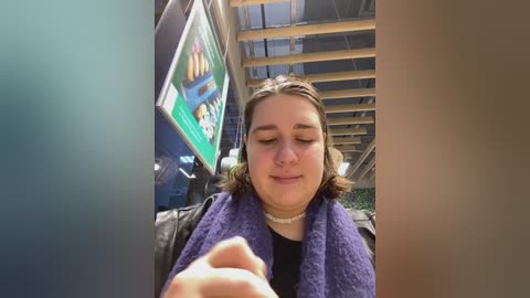 Media: Video of a young woman with shoulder-length brown hair, wearing a purple scarf and a black shirt, sitting on a bus with a green poster and wooden ceiling.