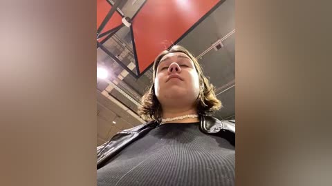 Media: Video of a plus-sized woman with short, wavy brown hair, wearing a black ribbed sweater and a black leather jacket, standing in a modern, industrial-style room with red and black accents.