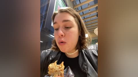 Media: Video of a young woman with shoulder-length brown hair, wearing a black leather jacket and choker, eating a sandwich in a modern, sunlit indoor space with exposed wooden beams.