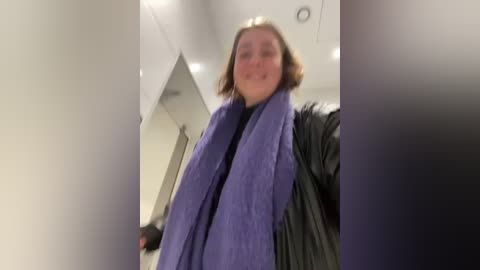 Media: Video of a woman with shoulder-length brown hair, wearing a purple scarf and black coat, walking down a dimly lit, modern hallway with white walls and a black coat rack.