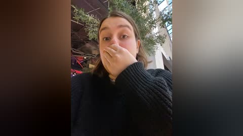 Media: Video of a young woman with light skin and brown hair, wearing a dark sweater, covering her mouth with her hand, in an indoor setting with greenery and architectural details.