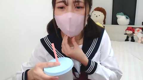 Media: A video of an Asian woman in a white sailor uniform with a pink mask, drinking from a blue and pink straw cup. The background features stuffed toys, including a white bunny and a red doll.