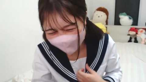 Media: Video of a young Asian girl with straight black hair and pale skin, wearing a white school uniform with a black collar and a pink surgical mask, crying in a white room with stuffed toys.