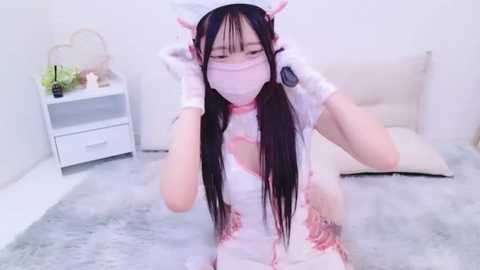 Media: Video of a young Asian woman with long black hair, wearing a pink nurse costume, white gloves, and mask, kneeling on a soft, light gray carpet in a minimalist, white-walled room.