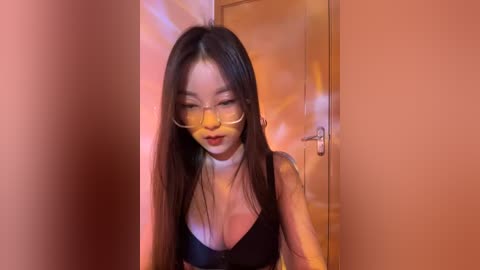 Media: A video of an East Asian woman with long black hair, wearing glasses, a black bra, and a choker, standing in a hallway with peach-colored walls.