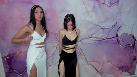 Media: Video of two women in white and black dresses with high slits, standing against a marbled purple and white background. One woman has long black hair, the other short black hair.