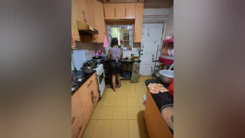 Media: Video of a small, cluttered kitchen with yellow tiles, wooden cabinets, and a person in a purple shirt and black skirt facing away. Stove, sink, and cluttered countertops with food items.