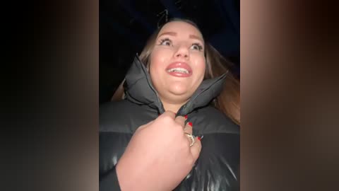 Media: A video of a woman with light skin, brown hair, and red lipstick, wearing a black puffy jacket, smiling and looking up, taken in a dimly lit room.