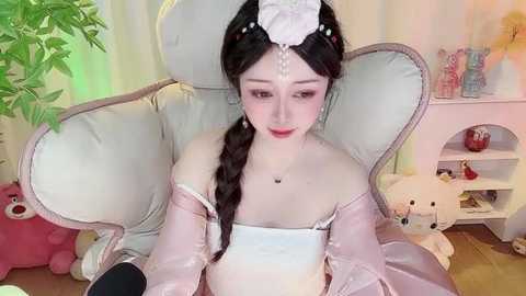 Media: Video of an Asian woman with fair skin, long braided hair, and a pink floral headpiece, wearing a white off-shoulder dress, seated in a pastel room with plush toys and a teddy bear.