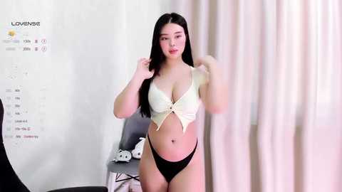 Media: A video of an East Asian woman with long black hair, fair skin, and a slim physique, wearing a white crop top and black thong, standing in a modern, white-walled room with a calendar, a chair, and a white curtain.