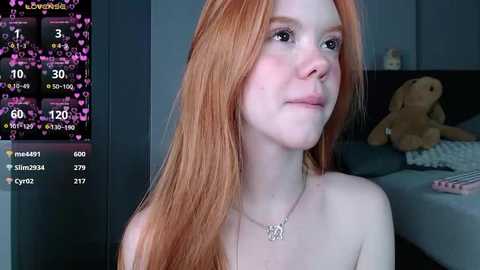 Media: Video of a fair-skinned, red-haired young woman with freckles, wearing a necklace, sitting indoors, surrounded by a virtual chat interface displaying hearts and numbers.