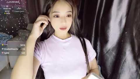 Media: Video of a young East Asian woman with long black hair, wearing a light pink t-shirt, seated against a black backdrop. She has a slender physique and is engaged in a livestream with audience reactions displayed.