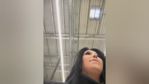 Media: Video of a woman with long black hair, wearing a black coat, looking upwards at a high ceiling with exposed pipes and industrial lighting.