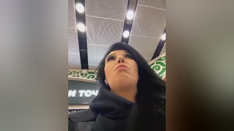 Media: Video of a woman with medium skin tone, wearing a black hijab and dark eyeliner, looking upward, in a modern, well-lit indoor setting with a green and white patterned ceiling.