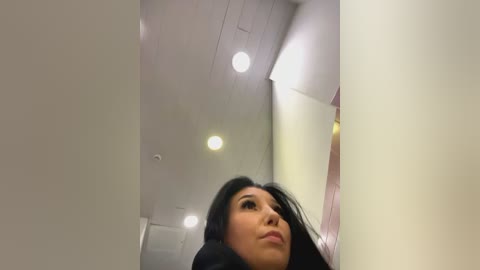 Media: A low-angle video of an Asian woman with long black hair and fair skin, wearing a light pink top, standing in a modern, well-lit indoor hallway with white walls and recessed ceiling lights.