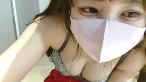 Media: Video of a young woman with light skin, brown hair, and brown eyes, wearing a gray lace bra and a white face mask, taken from a high angle, showing cleavage.